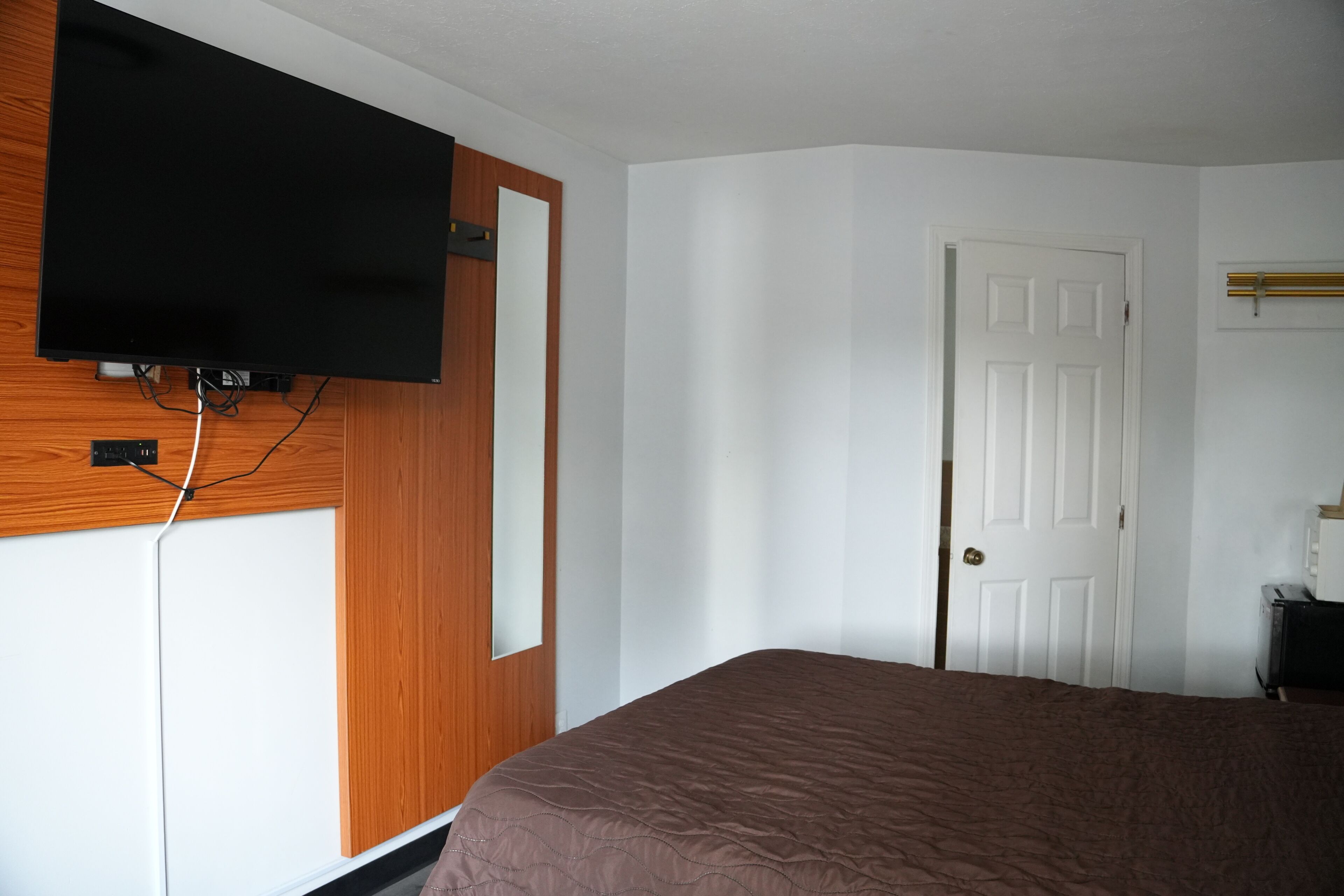 A hotel room with a bed, a television mounted on a wooden panel, a white door, and a small desk with a lamp.