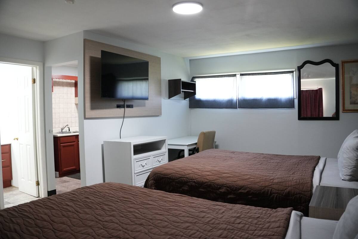 Una camera d'hotel con due letti, una scrivania, una televisione, una finestra con tende e uno specchio.