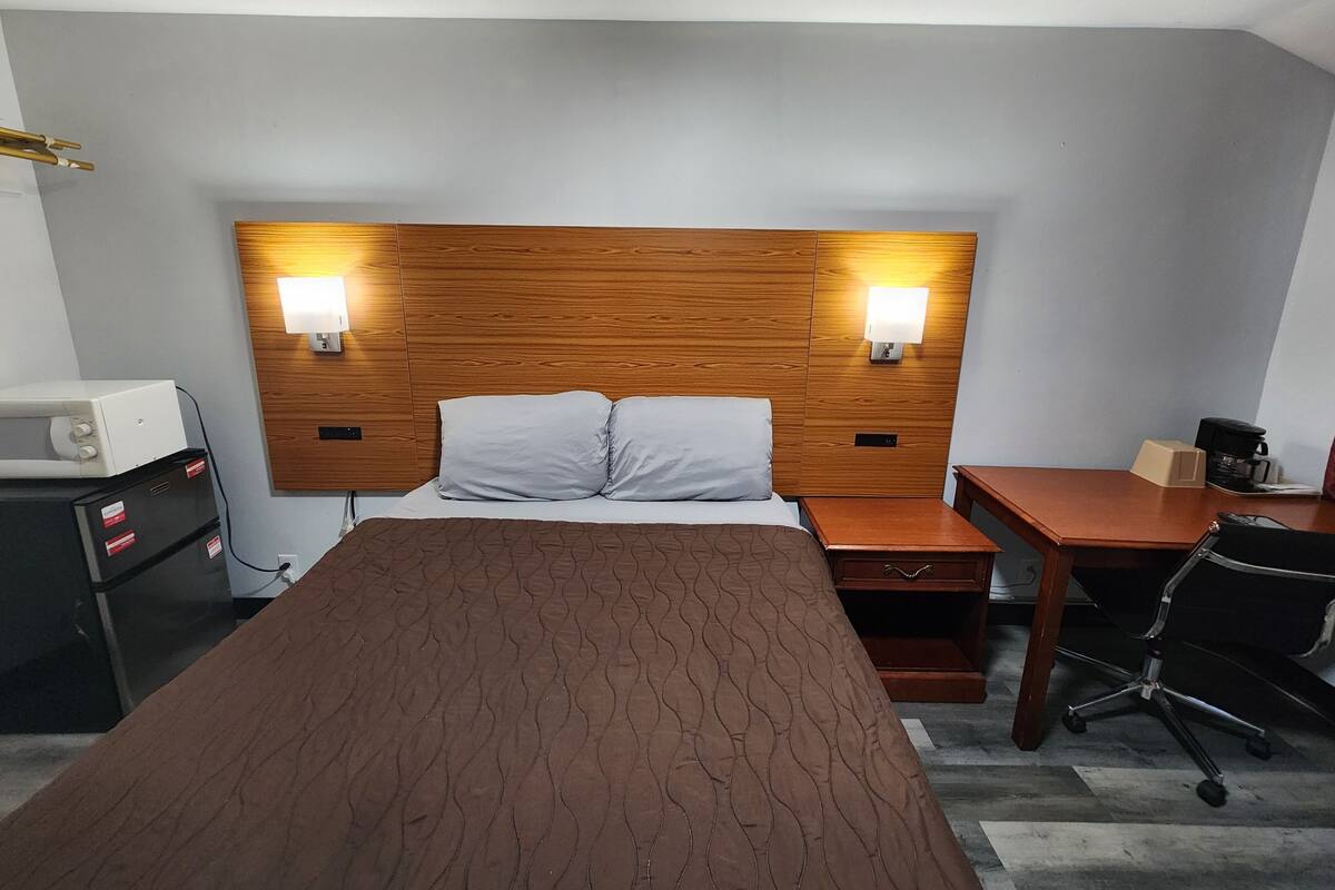 Una camera d'albergo con una testiera in legno, due lampade sui comodini, un letto con copriletto marrone, una scrivania con una sedia e una macchina per il caffè.