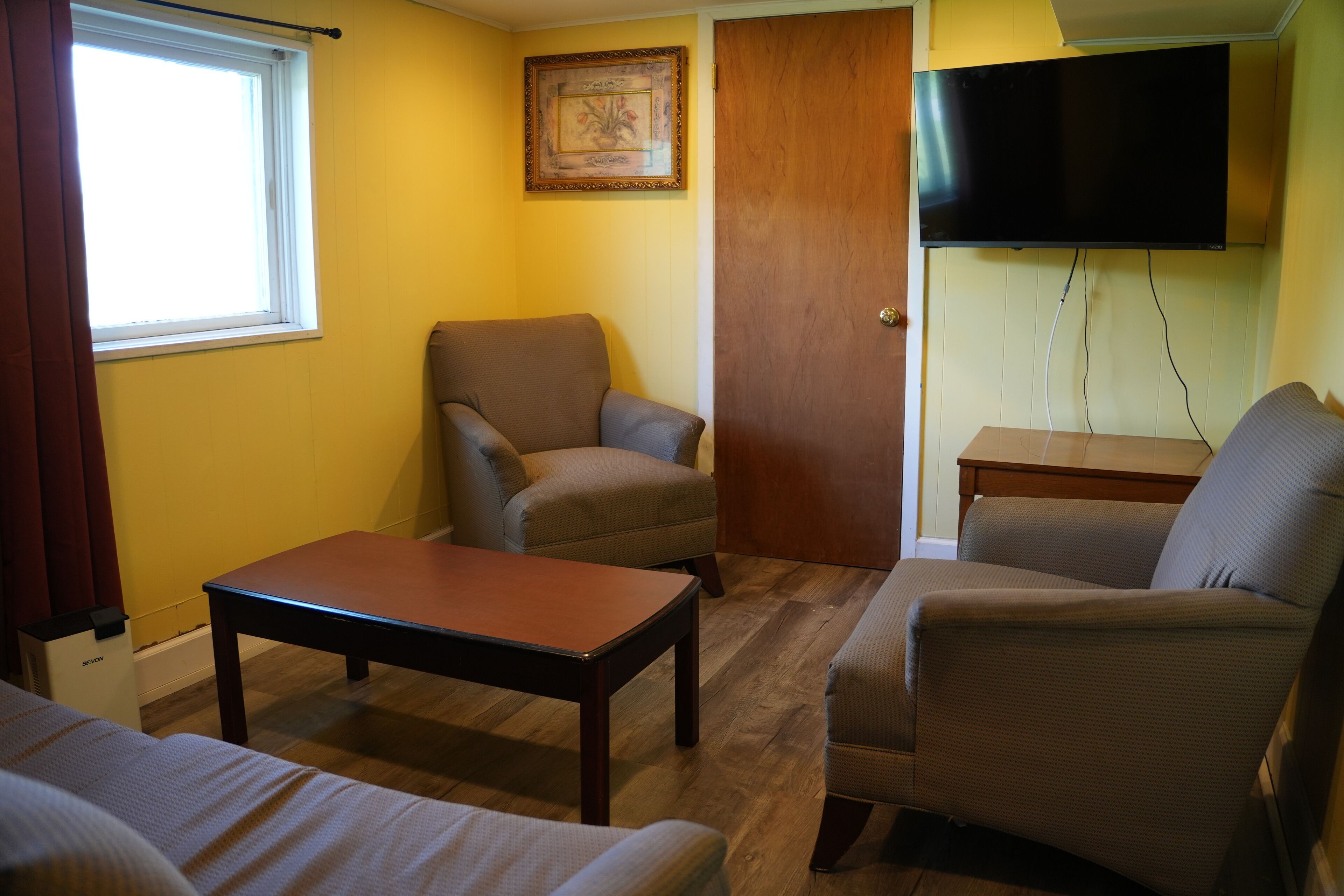 A room with a wooden floor, a sofa, a chair, a table, a TV mounted on the wall, and a door.