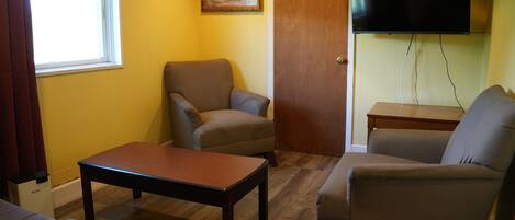 A room with a wooden floor, a sofa, a chair, a table, a TV mounted on the wall, and a door.