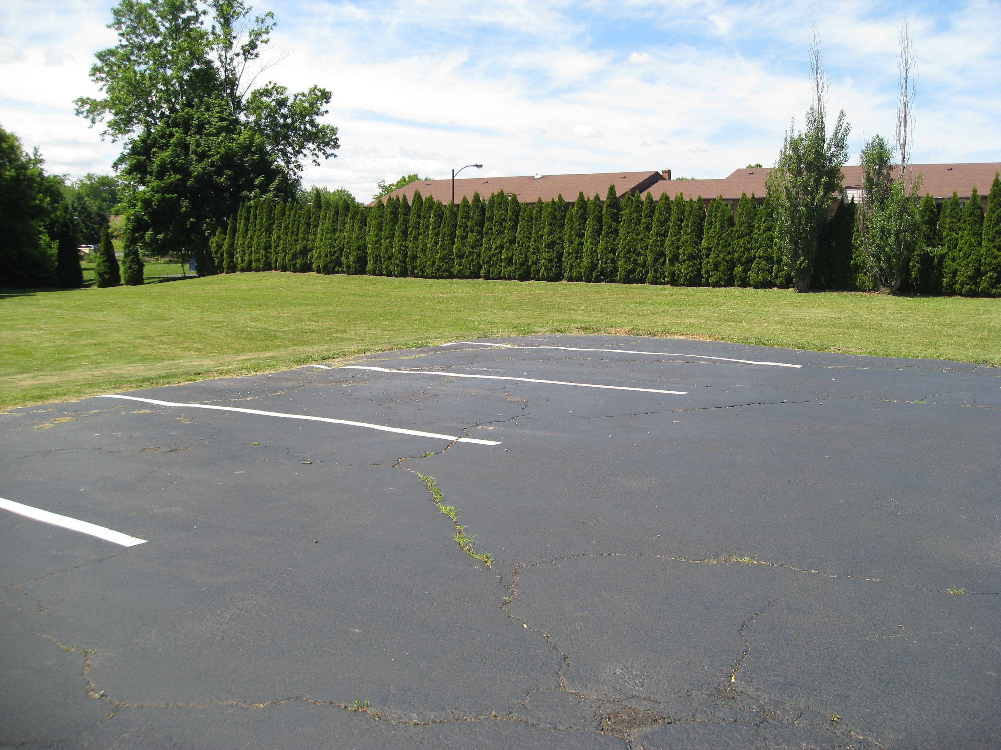 A parking lot with visible cracks and faded lines, surrounded by a well-maintained lawn and a row of evergreen trees.