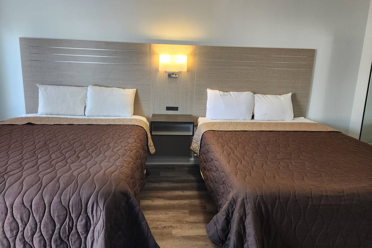 Una camera d'albergo con due letti, ciascuno con una coperta trapuntata marrone e cuscini bianchi. Tra i letti c'è un comodino con una lampada.