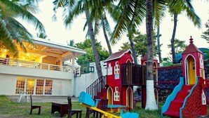 Un parque infantil con un sube y baja, tobogán y estructuras para escalar, rodeado de palmeras y un edificio con grandes ventanales.