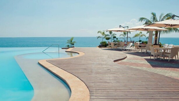 Terraza de madera con piscina que da al mar, sillas de descanso y sombrillas.
