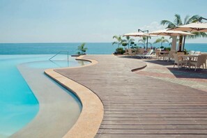Een houten terras met zwembad dat uitkomt op de zee, ligbedden en parasols.