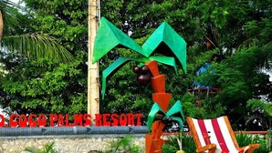 Letrero del Resort Coco Palms con una escultura estilizada de una palmera y una silla de playa a rayas.