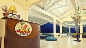 Una recepción con el logotipo de Coco Palms Resort, un vestíbulo amplio con techo alto y una zona de estar con sillas y un sillón de peluche azul.
