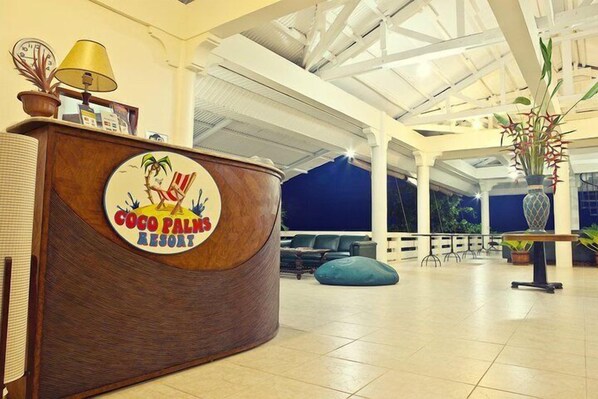 A reception desk with a logo for Coco Palms Resort, a spacious lobby with a high ceiling, and a seating area with chairs and a blue bean bag.