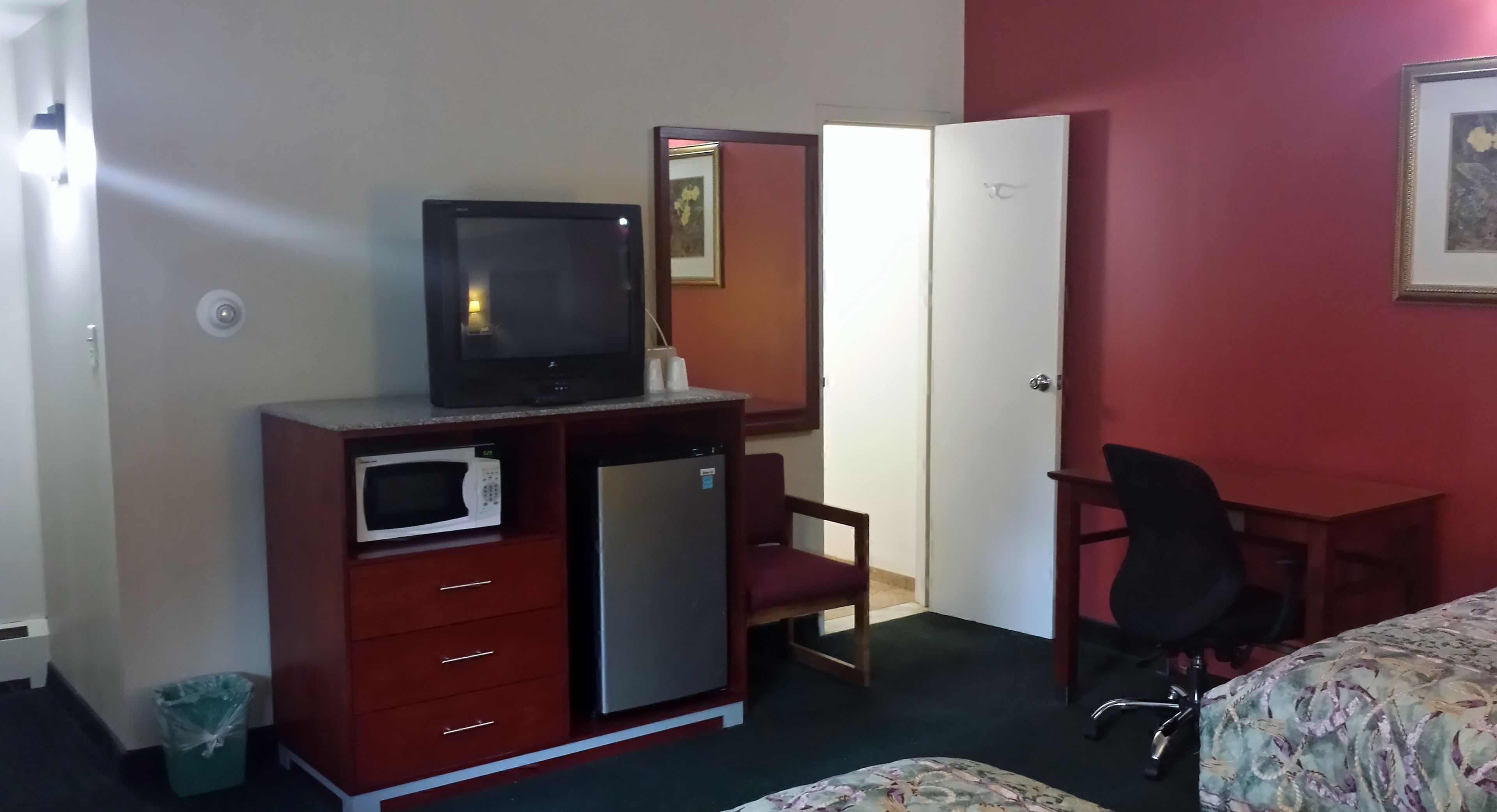 Quarto de hotel com uma cama, uma escrivaninha com cadeira, TV, micro-ondas, frigorífico e lixeira.