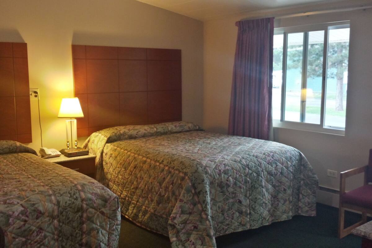 A hotel room with two beds, a nightstand, a lamp, a chair, and a window with curtains.