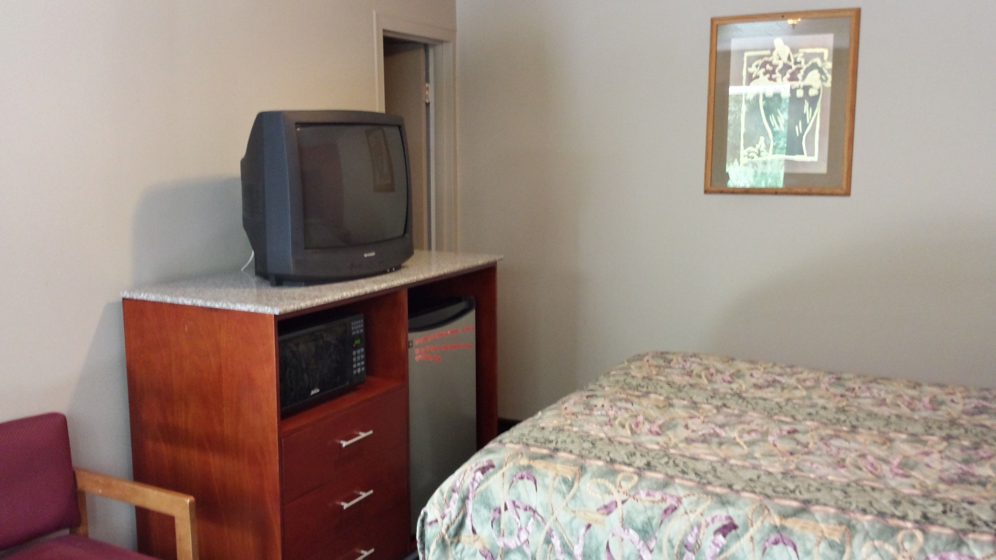 A bedroom with a bed, a wooden dresser with a television on top, a microwave, and a framed picture on the wall.