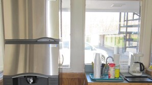 A kitchen with a stainless steel refrigerator, a water dispenser, wooden cabinets, and a window with a view of the street. - Budget Inn Fairport (Fairport)