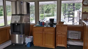 A kitchen area with a stainless steel refrigerator, a coffee machine, and a window with a view of the parking lot and trees. - Budget Inn Fairport (Fairport)