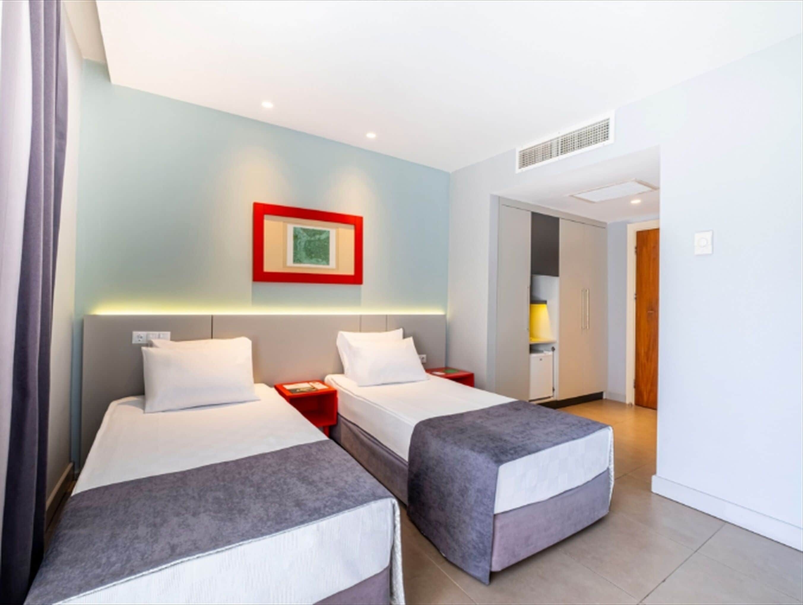 A hotel room with two beds, a red-framed picture on the wall, a bedside table with a lamp, and a wardrobe.