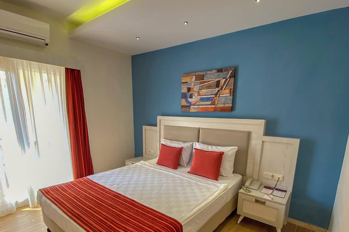 A bedroom with a bed, a nightstand, a painting on the wall, and a window with curtains.