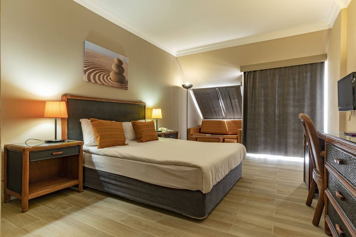 A hotel room with a bed, bedside tables, a desk, a chair, a television, and a large window with curtains.