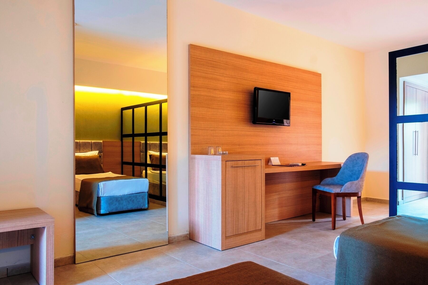 Una habitación de hotel moderna con un escritorio de madera, un televisor montado en la pared, una silla azul y un espejo grande que refleja la habitación.