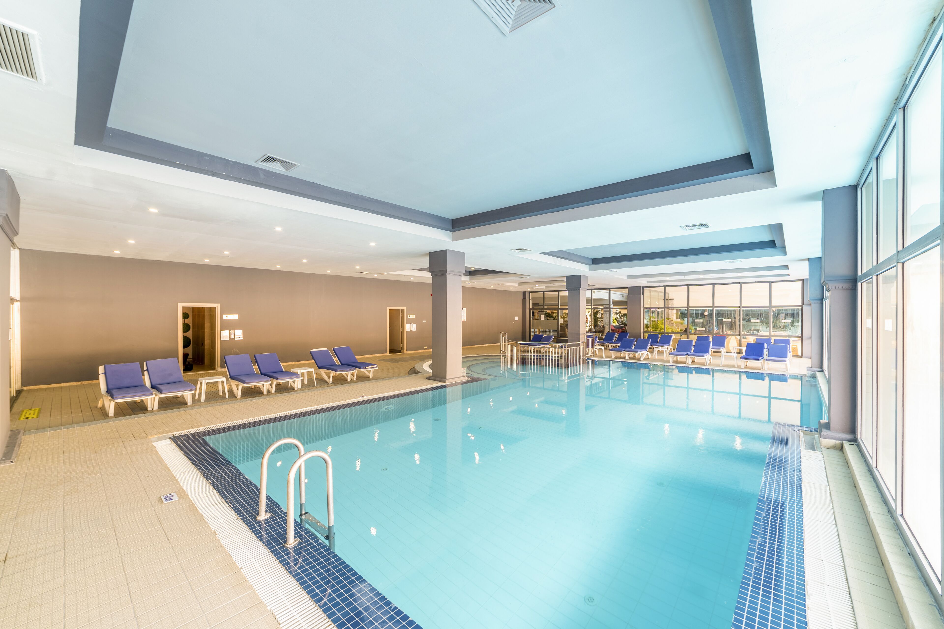 An indoor swimming pool with blue and white tiled floors, a clear pool edge, and blue lounge chairs arranged around the pool.