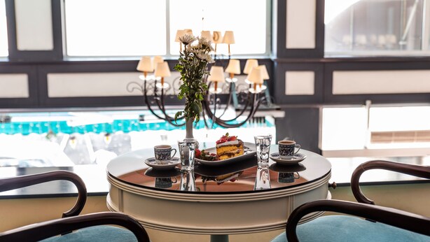 A round table with a cake and tea set, surrounded by chairs with blue upholstery, in a room with large windows and a view of parked cars. - Viva Deluxe Hotel (Istanbul)