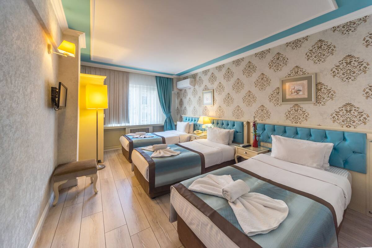 A hotel room with two beds, a bedside table with a lamp, a window with curtains, and a wall with a decorative pattern.