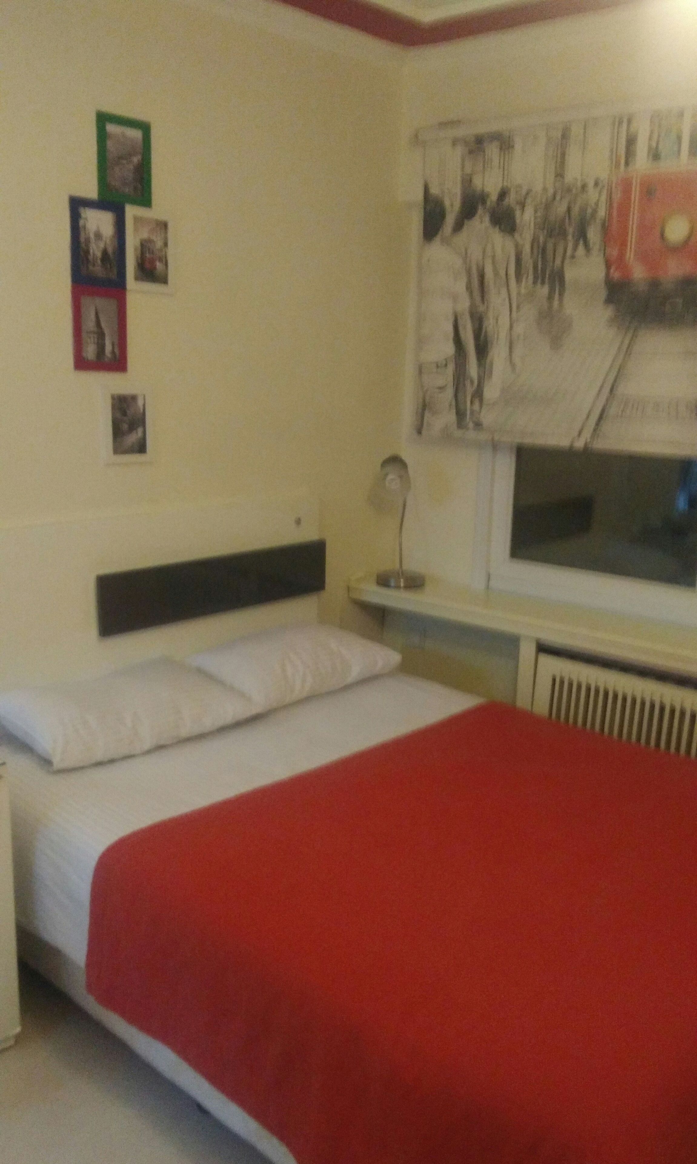 Een slaapkamer met een bed, nachtkastje, lamp, raam en een muur met foto’s.