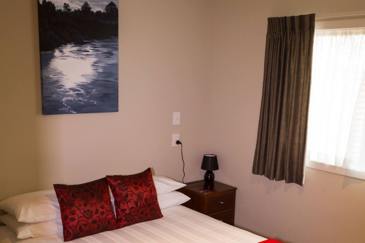 A neatly made bed with a red and white bedspread, a single red patterned pillow, a bedside table with a lamp, and a painting of a landscape on the wall.