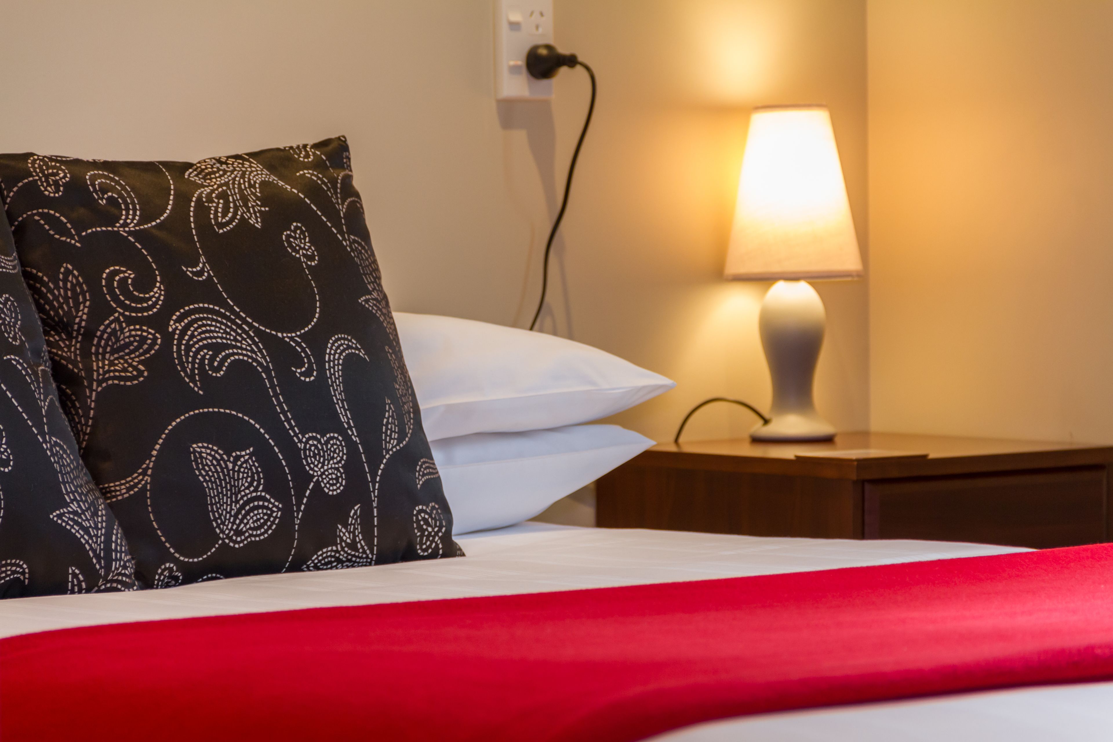 Un lit bien fait, recouvert d’une couverture rouge, avec un oreiller à motifs noir et blanc et un oreiller blanc. Une lampe de chevet est posée sur une table de chevet en bois.