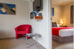 Una habitación de hotel con un sillón rojo, una mesita pequeña, una cama con una colcha roja, un televisor montado en la pared y un cuadro en la pared.