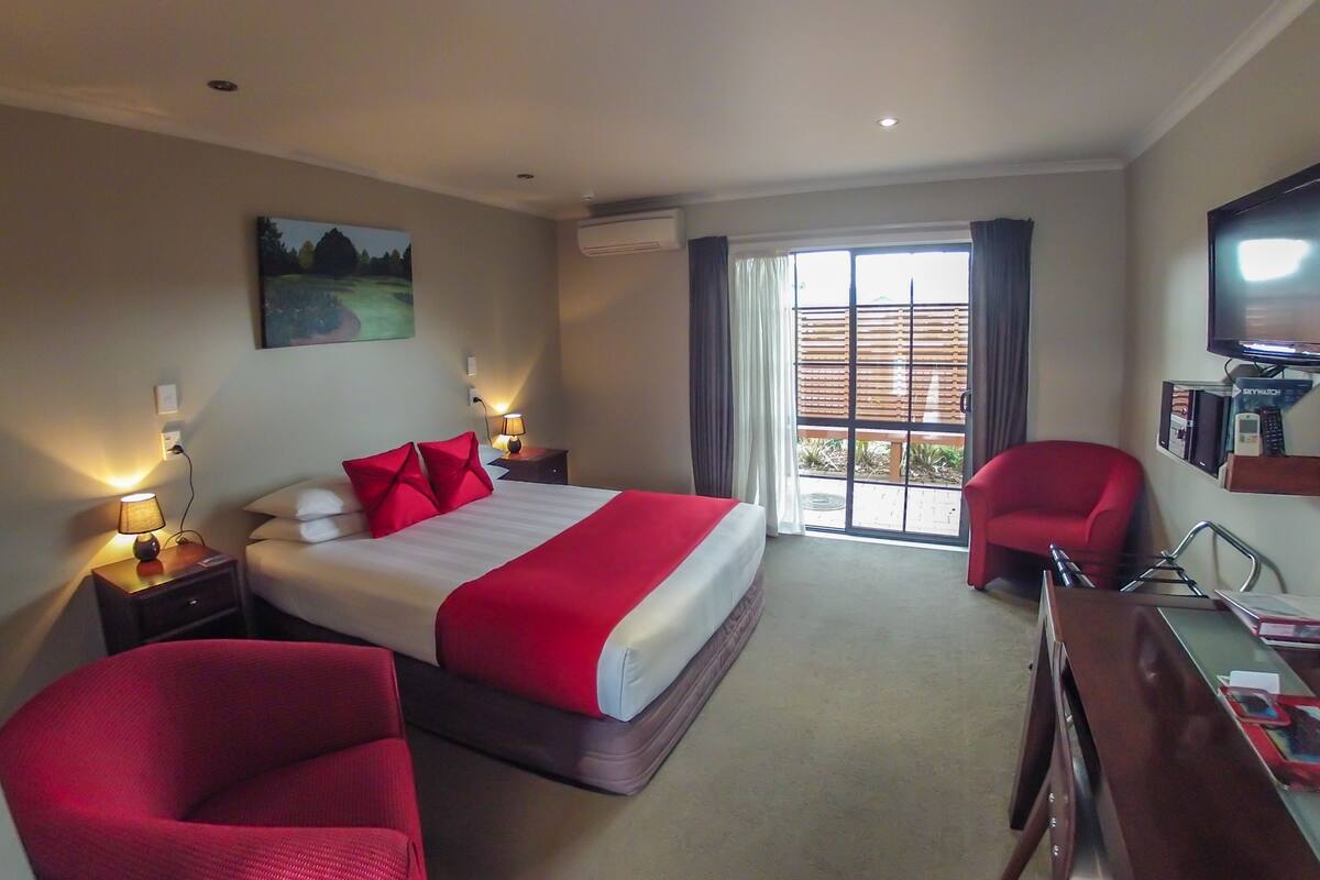 A hotel room with a bed, two red chairs, a desk, a television, and a sliding glass door to a balcony.