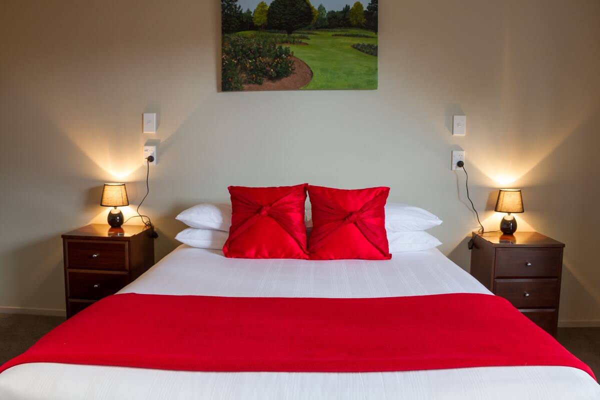 A neatly made bed with white and red bedding, two bedside tables with lamps, and a painting of a landscape on the wall.
