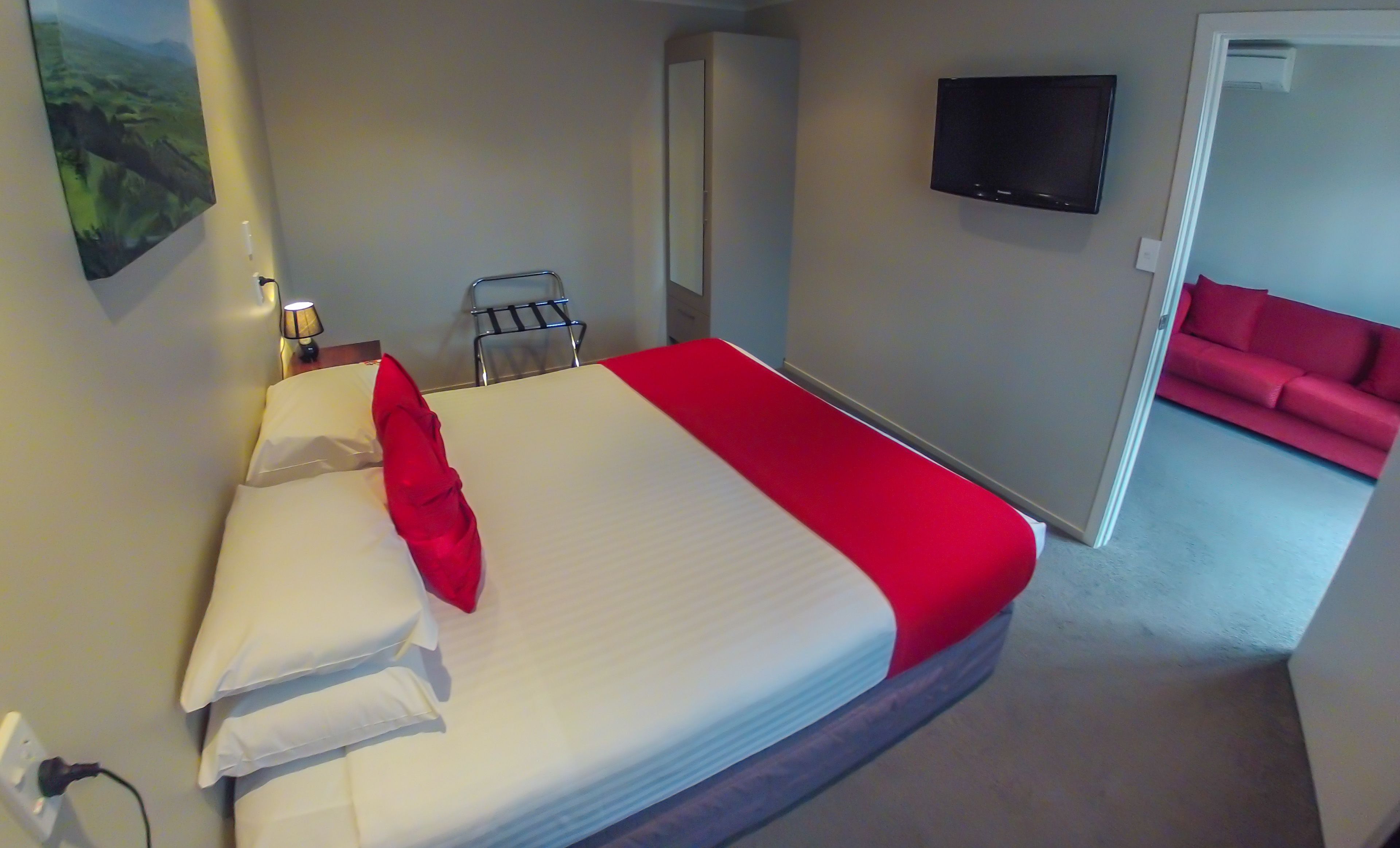Een hotelkamer met een bed, een aan de muur bevestigde televisie, een rode bank in de aangrenzende zithoek, en een landschapsschilderij aan de wand.