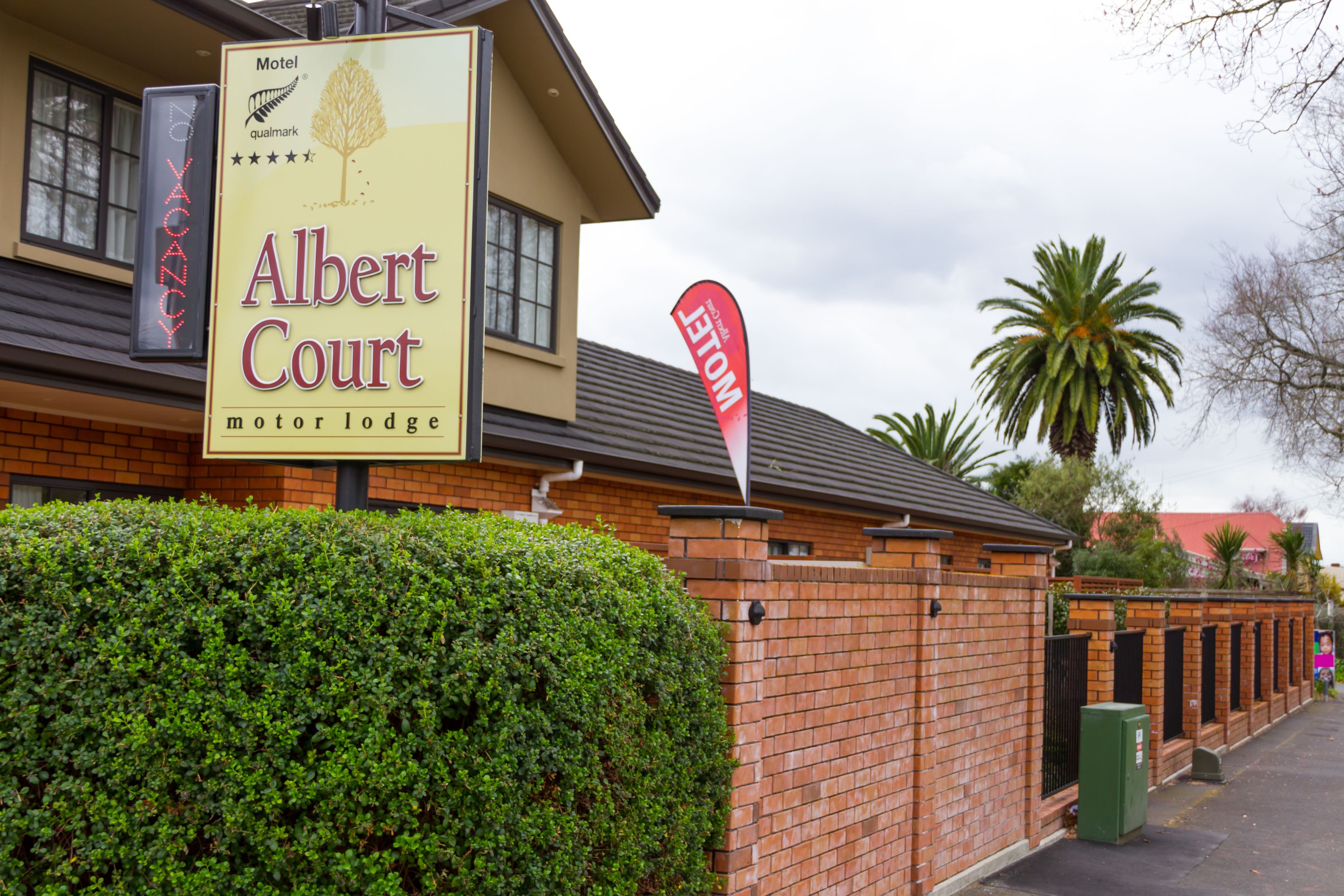 A motel named Albert Court with a signboard, a brick wall, and a well-maintained hedge.