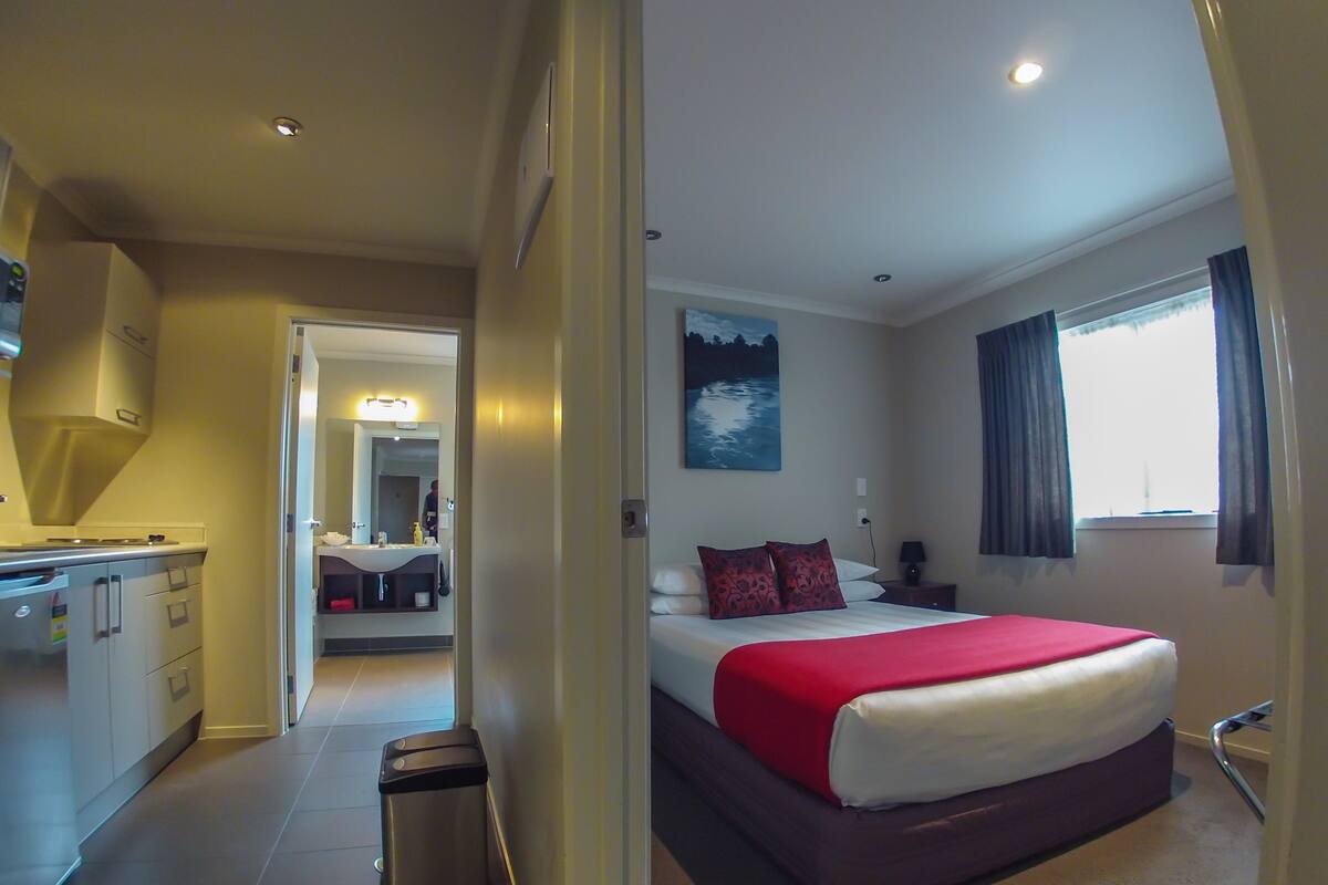 A hotel room with a bed, a nightstand, a window with curtains, a painting on the wall, and a kitchenette with a sink and cabinets.