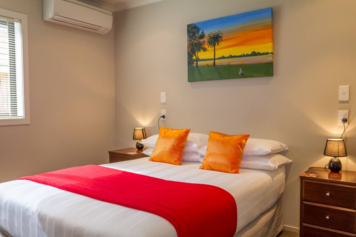 A hotel room with a bed, two nightstands, a painting of a sunset, and an air conditioner on the wall.