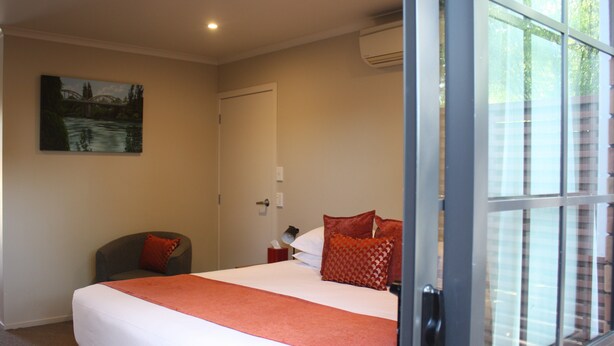 A hotel room with a bed, a chair, a painting on the wall, and a window with a view of buildings. - Albert Court Motor Lodge (Hamilton)