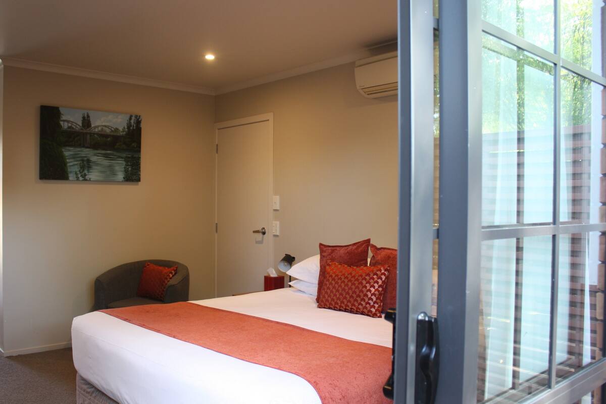 A hotel room with a bed, a chair, a painting on the wall, and a window with a view of buildings.