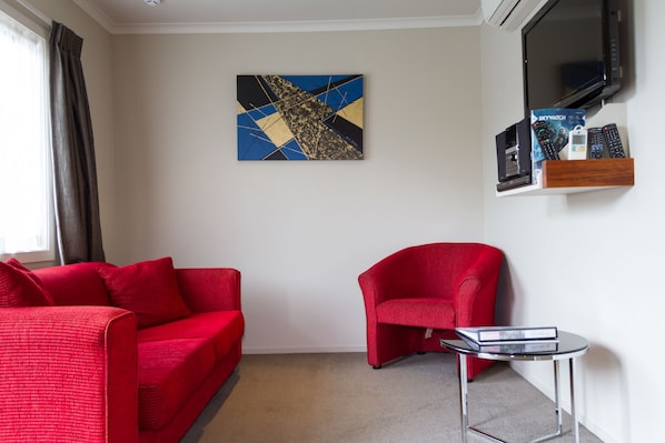 Un salon moderne avec un canapé rouge, un fauteuil rouge et une petite table. Un téléviseur à écran plat est fixé au mur, et un tableau est accroché au-dessus.