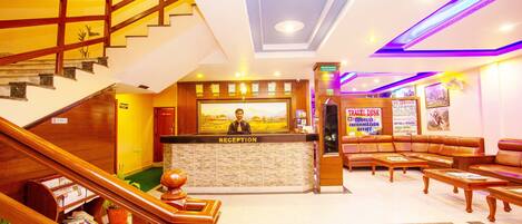 A hotel reception area with a reception desk, a staircase, a seating area with sofas, and a reception desk with a framed picture.