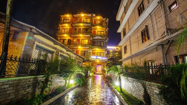 Un hotel de varios pisos con una entrada central, rodeado por un estrecho callejón con paredes de ladrillo y vegetación.