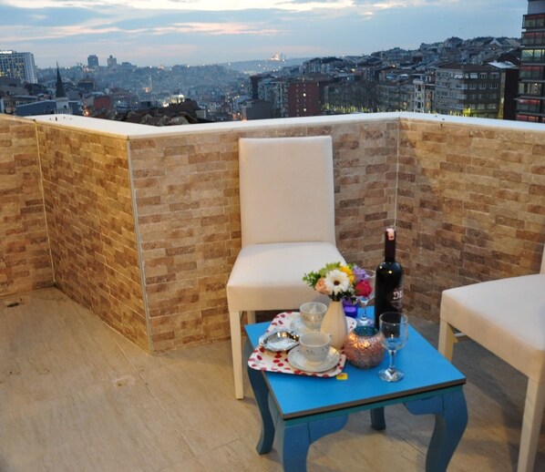 Eine Dachterrasse mit Backsteinmauer, einem weißen Stuhl, einem blauen Tisch gedeckt mit einem Blumenstrauß, einer Weinflasche und einem Glas.