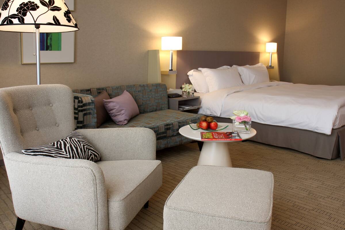 Una habitación de hotel con una cama, dos sillones, un sofá, una mesita con fruta y una revista, y dos lámparas de noche.