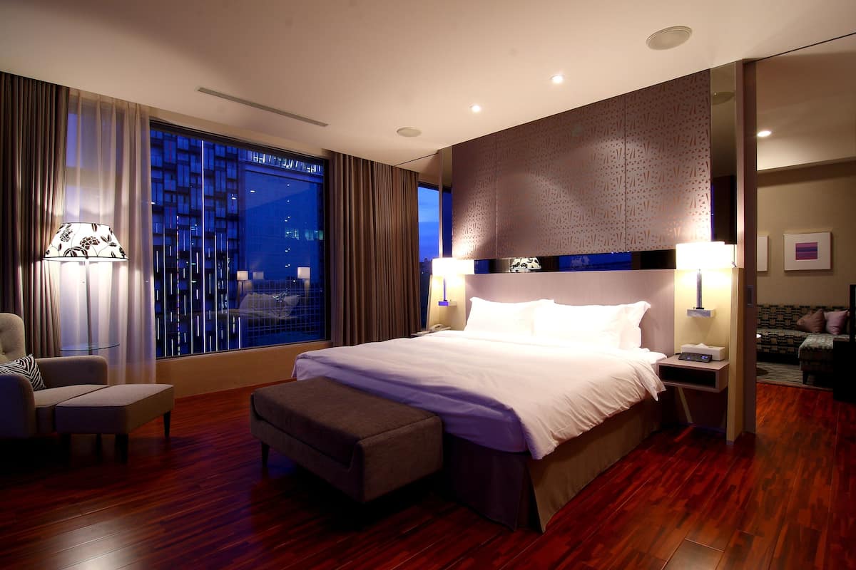 Una habitación de hotel moderna con una cama grande, una zona de estar con una silla y un reposapiés, una mesita de noche con una lámpara, y vistas a la ciudad de noche.