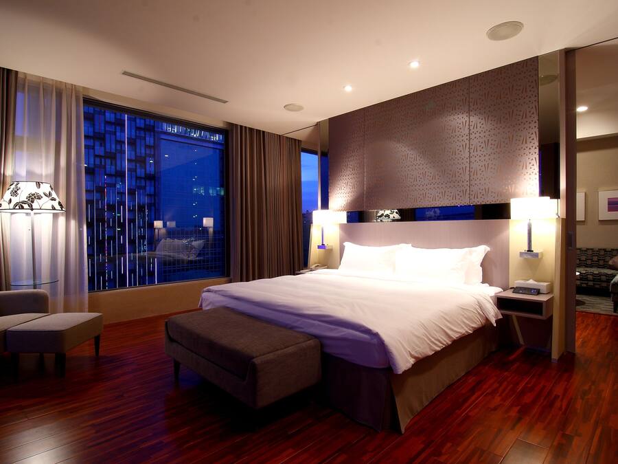 A modern hotel room with a large bed, a sitting area with a chair and ottoman, a nightstand with a lamp, and a view of the city at night.
