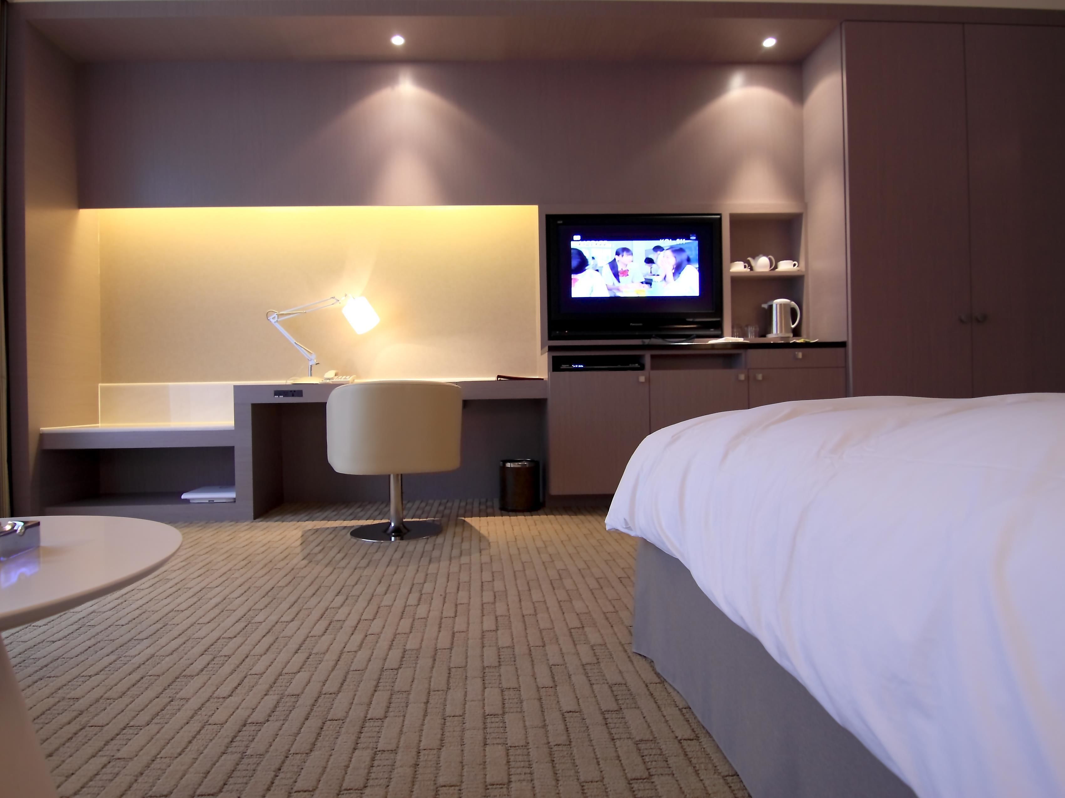 A hotel room with a bed, a desk with a lamp, a television, and a minibar.