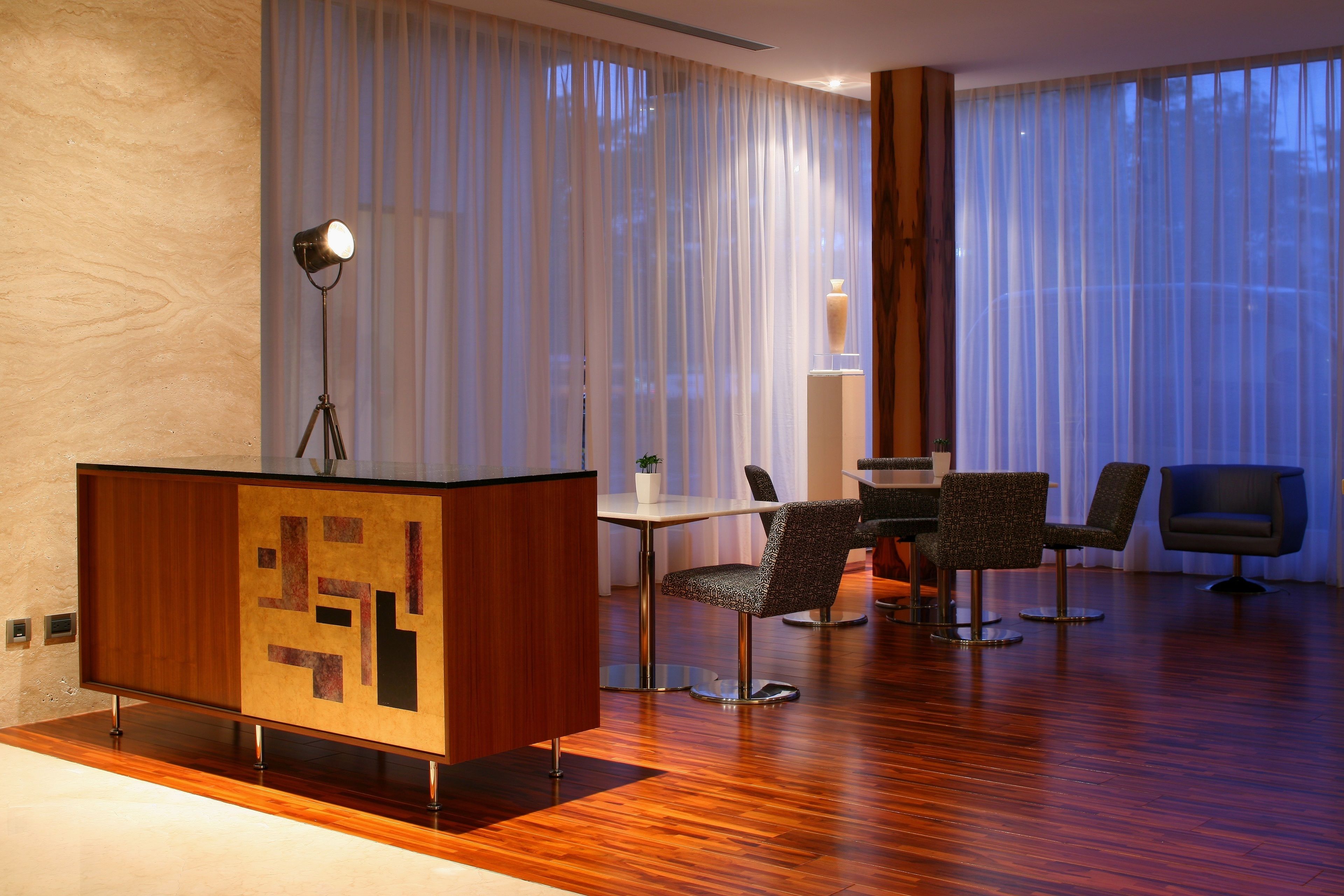 Een moderne hotelkamer met een houten vloer, een bureau met lamp en een zithoek met stoelen.