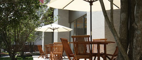 Mesa al aire libre con sillas de madera y sombrillas grandes, un árbol y un edificio en el fondo.