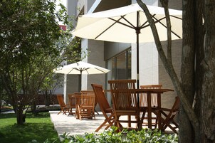 Outdoor seating with wooden chairs and large umbrellas, a tree, and a building in the background.