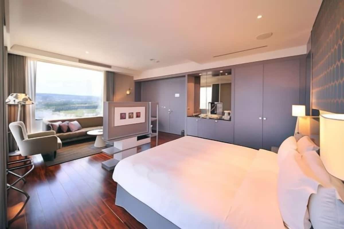 Una habitación de hotel moderna con una cama grande, una zona de estar con sofá y sillones, un televisor de pantalla plana y un ventanal con vistas.