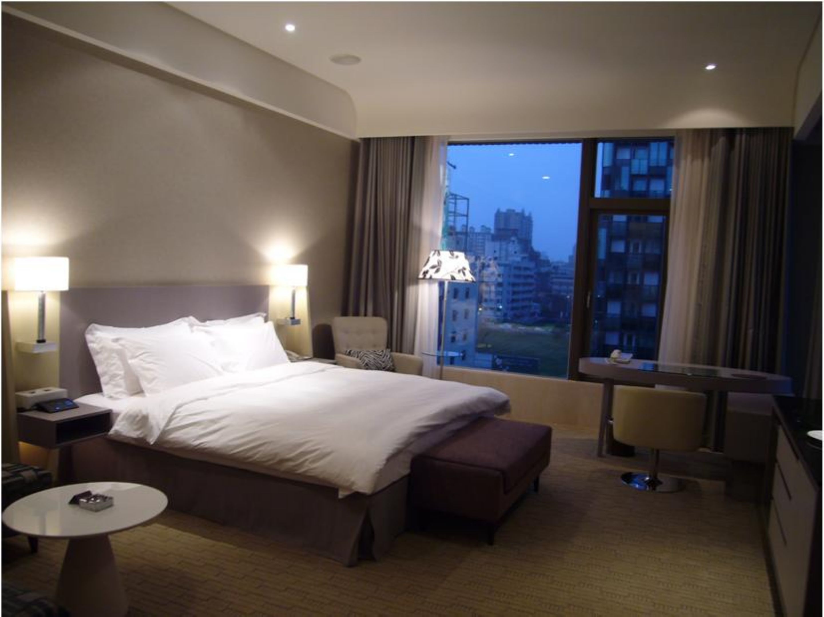 Habitación de hotel con una cama grande, dos lámparas de noche, una mesita pequeña, una silla y vistas a la ciudad desde la ventana.